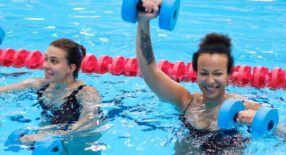 twee lachende vrouwen die aquarobics doen in hget zwembad met dumbells van foam